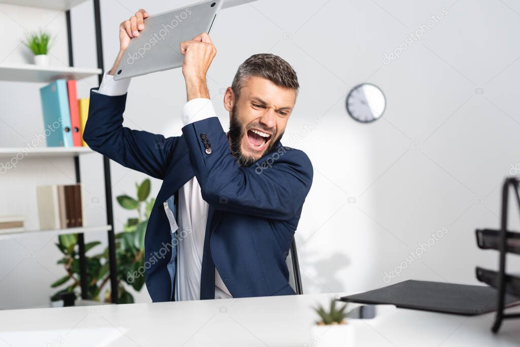 Nervous businessman holding laptop near stationery on blurred foreground in office 