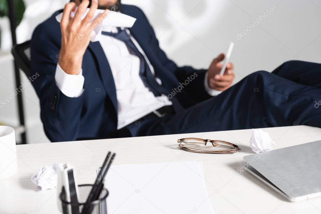 Eyeglasses and laptop on table near businessman holding clumped paper on blurred background 