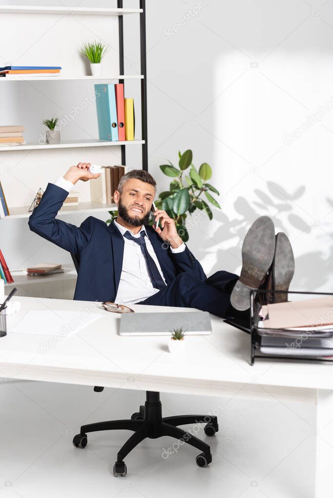 Angry businessman holding clumped paper while talking on smartphone in office 