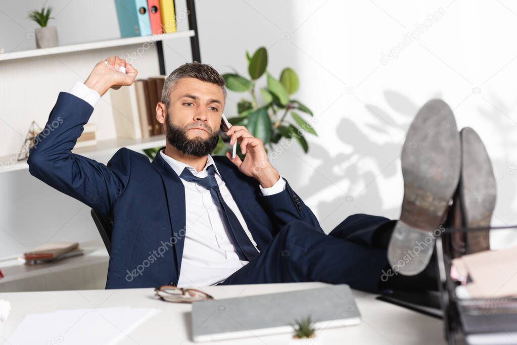 Businessman holding clumped paper while talking on smartphone near laptop and papers on blurred foreground 