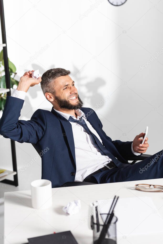 Angry businessman holding clumped paper and smartphone near stationery on blurred foreground in office 