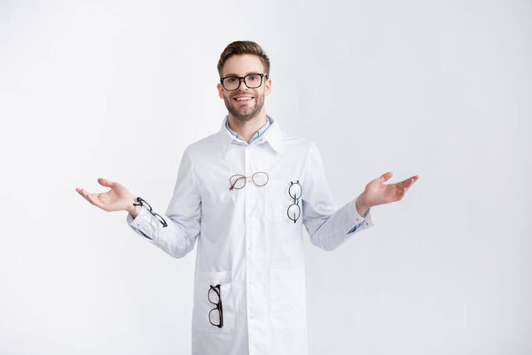 Front view of smiling ophthalmologist with open arms wearing white coat with hanging pairs of eyeglasses isolated on white