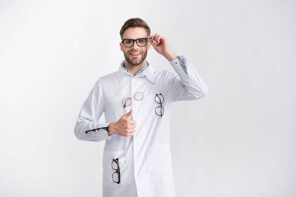 Front view of smiling ophthalmologist with thumb up, wearing white coat with hanging pairs of eyeglasses isolated on white