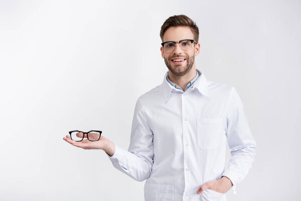 Вид спереди улыбающегося врача с рукой в кармане, держащего очки на ладони, при взгляде на камеру изолированную на белом
