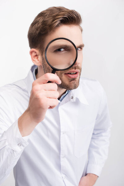 Curious ophthalmologist in white coat looking through magnifying glass isolated on white