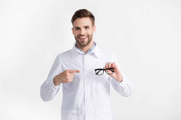 Вид спереди счастливого врача, указывающего пальцем на очки, при этом глядя на камеру, изолированную от белого