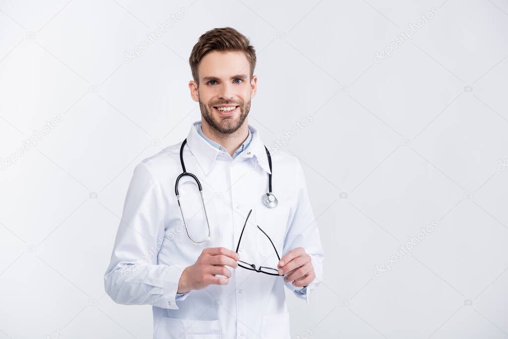 Front view of happy ophthalmologist in white coat holding eyeglasses, while looking at camera isolated on white