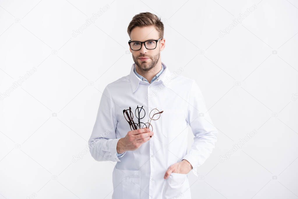 Front view of young adult ophthalmologist with hand in pocket holding several eyeglasses isolated in white