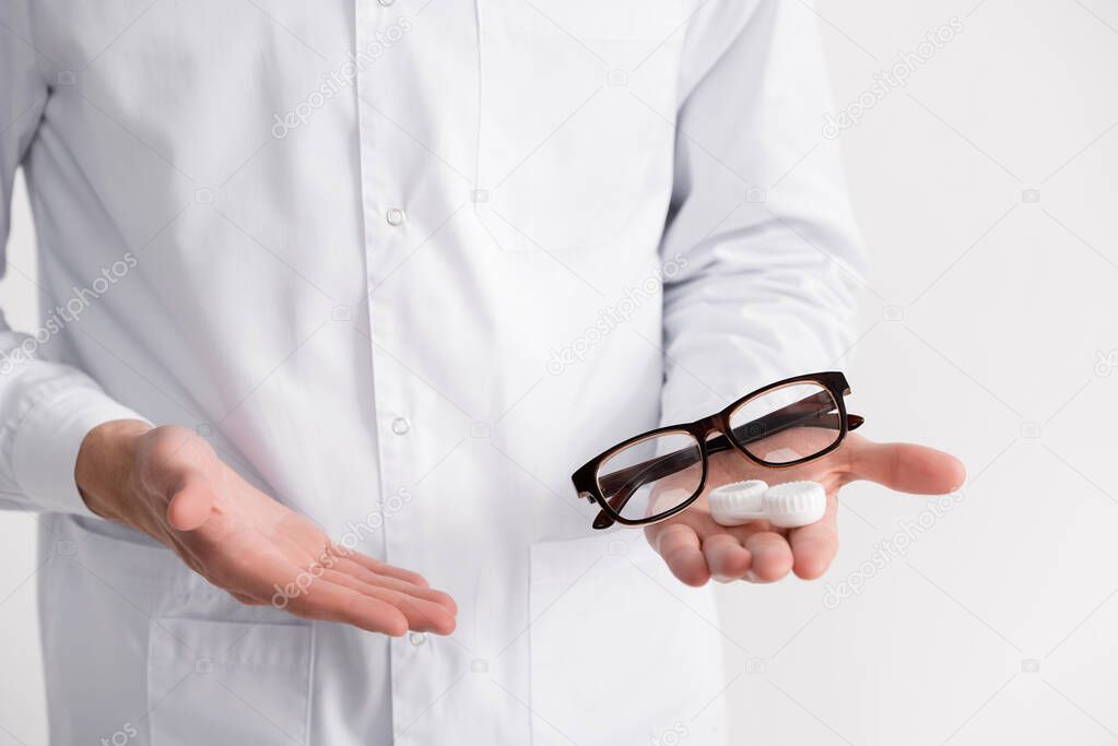 Cropped view of ophthalmologist pointing with hand at eyeglasses and lenses container on palm isolated on white
