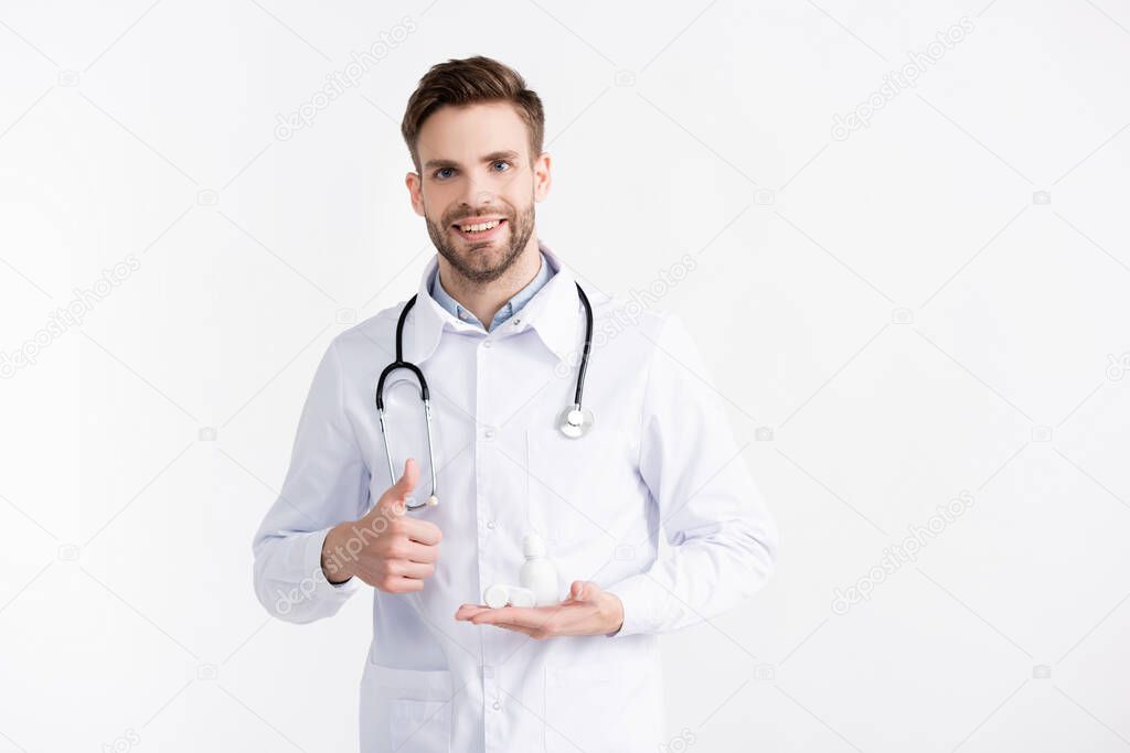 Front view of smiling ophthalmologist with thumb up, holding lenses container and plastic bottle isolated on white