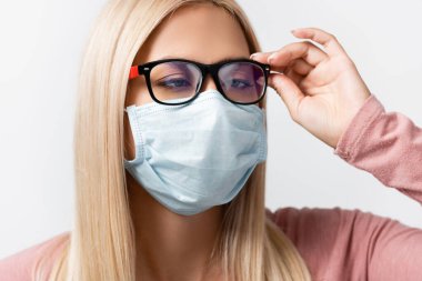 Squint woman in medical mask touching eyeglasses isolated on grey clipart