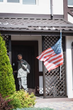 Serious military man standing near house door and looking away near bushes and american flag clipart