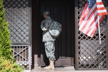 Back view of military man with backpack standing near house door and american flag clipart