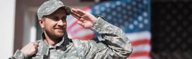 Gülümseyen asker arka planda bulanık Amerikan bayrağıyla selamlıyor.