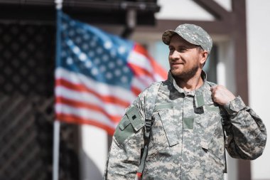 Kamuflajlı gülümseyen eski asker arka planda bulanık Amerikan bayrağıyla yüzünü çeviriyor.