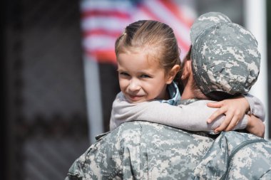 Arkasında bulanık Amerikan bayrağıyla gülümseyen kızına sarılan askerin arkası.