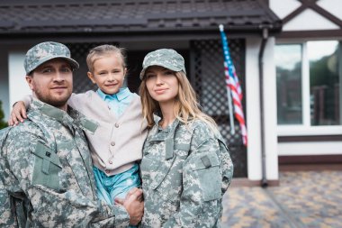Smiling father and mother in military uniforms lifting daughter and looking at camera on blurred background clipart