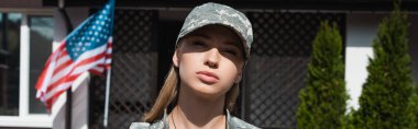 Ciddi bir asker kadın, Amerikan bayrağı ve pankartıyla evinin yakınındaki kameraya bakıyor.