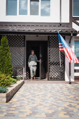 Military servicewoman standing in doorway near american flag and bushes clipart