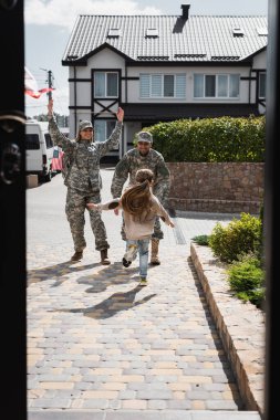 Daughter running to happy father and mother in military uniforms on street near house on blurred foreground clipart