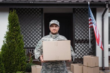 Front view of military serviceman with cardboard box standing near house with american flag clipart