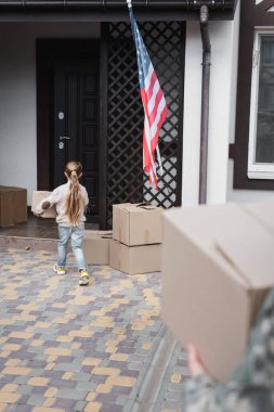 Little girl carrying cardboard box to house on blurred foreground clipart