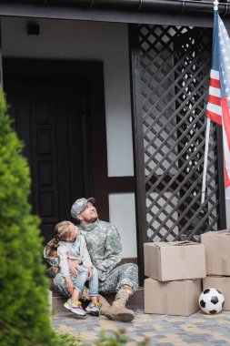 Askeri üniformalı baba eşikte oturmuş Amerikan bayrağına bakarken kızını kucaklıyor. 