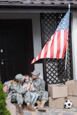 Kızı kucaklayan asker ebeveynler, karton kutuların yanında eşikte otururken ve ön planda bulanık Amerikan bayrağı