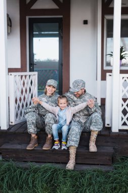 Happy daughter with outstretched hands sitting near mother and father in military uniforms on wooden threshold clipart