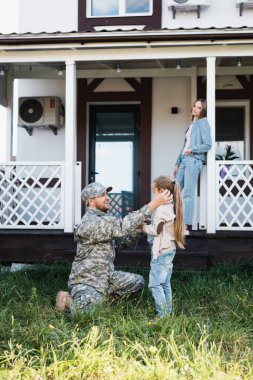 Military man sitting on knee and putting flower behind girl ear on backyard, with blurred woman and house on background clipart