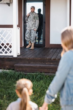 Happy military serviceman standing near back door on threshold with blurred woman and girl on foreground clipart