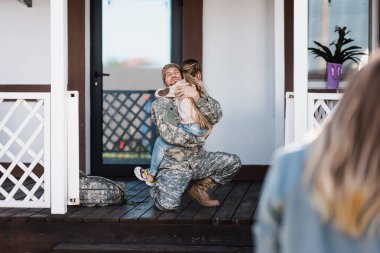 Smiling military service man embracing daughter, while sitting on knee on threshold with blurred woman on foreground clipart