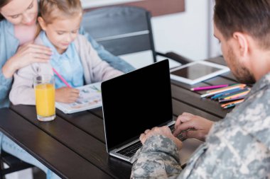 Askeri üniformalı bir adam boş ekranla dizüstü bilgisayarda yazarken arka planda bulanık kadın ve kızla masada oturuyor.
