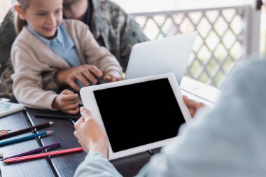 Elinde boş ekranlı dijital tablet tutan kadın. Arka planda bulanık asker ve kızı var.