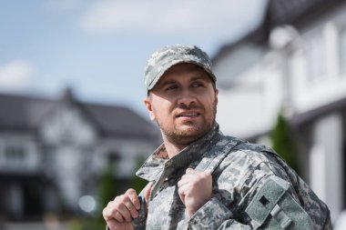 Confident military serviceman looking away, holding backpack shoulder straps with blurred neighborhood on background clipart