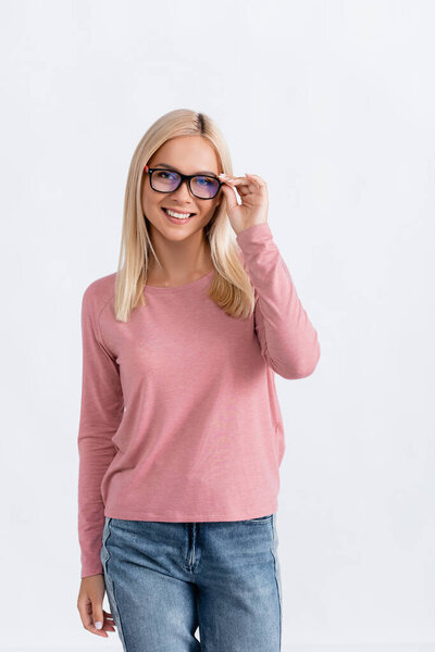 Smiling blonde woman in jeans and pink long sleeve holding eyeglasses frame, while looking at camera isolated on white