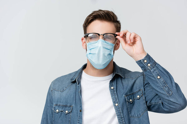 Young man in medical mask touching misted eyeglasses isolated on grey