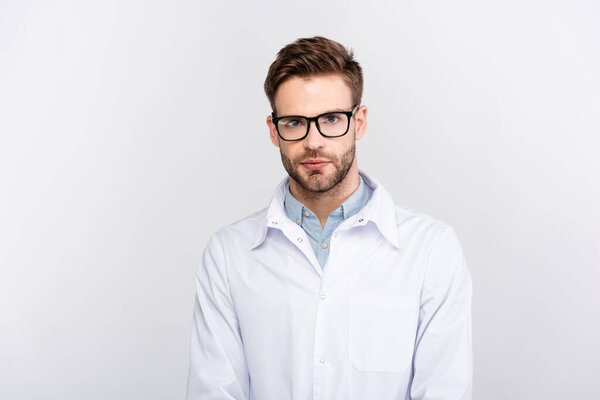 Young doctor in white coat and eyeglasses looking at camera isolated on grey 