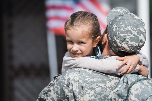 Back view of military man hugging smiling daughter with blurred american flag on background