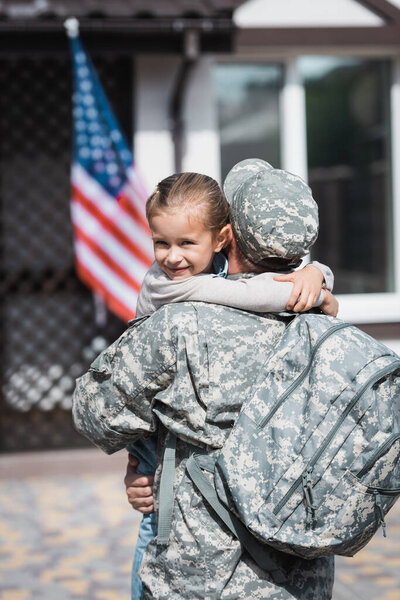 Back view of military man with backpack hugging daughter with blurred american flag and house on background
