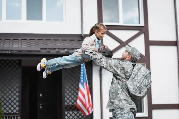 Военный Поднимает Улыбающуюся Дочь Воздухе Рядом Домом Американским Флагом — стоковое фото