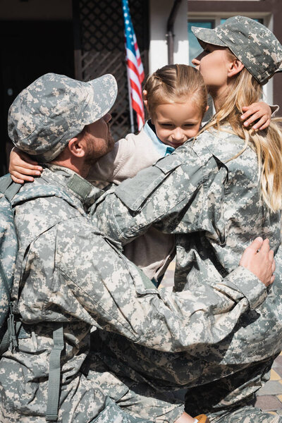 Upset daughter hugging mother and father in military uniforms near house with american flag