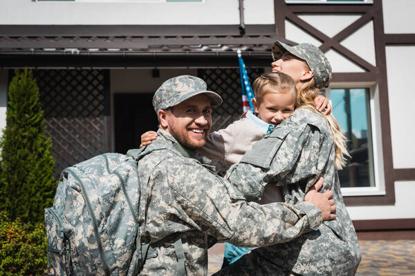 Smiling daughter hugging father and mother in military uniforms near house with american flag
