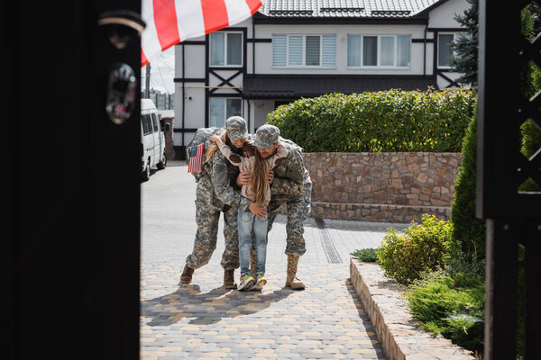 Daughter hugging mother and father in military uniforms on street near house on blurred foreground