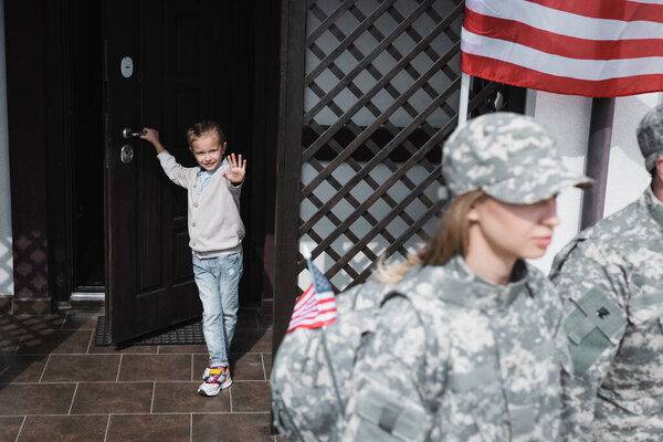 Smiling daughter with waving hand standing in doorway near american flag with blurred mother and father in military uniforms on foreground