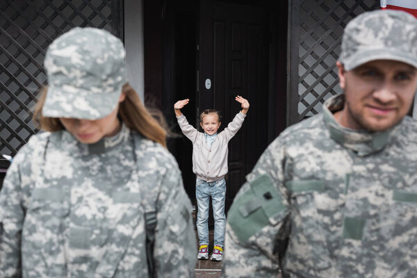 Smiling girl with waving hands standing near house door with blurred man and woman in military uniforms on foreground