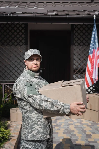 Militar Sosteniendo Caja Cartón Mientras Mira Cámara Cerca Casa Bandera — Foto de Stock