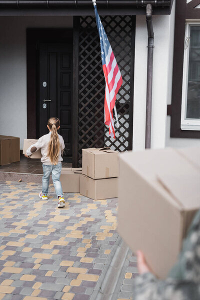 Little girl carrying cardboard box to house on blurred foreground