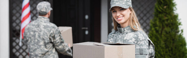 Happy military servicewoman holding cardboard box and looking at camera with blurred man on background, banner