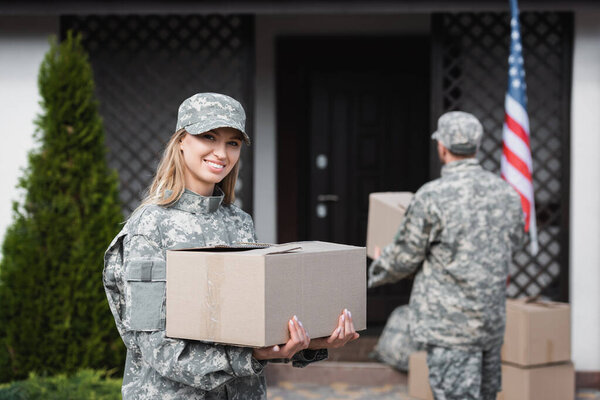 Smiling woman in camouflage holding cardboard box and looking at camera with blurred  military man on background